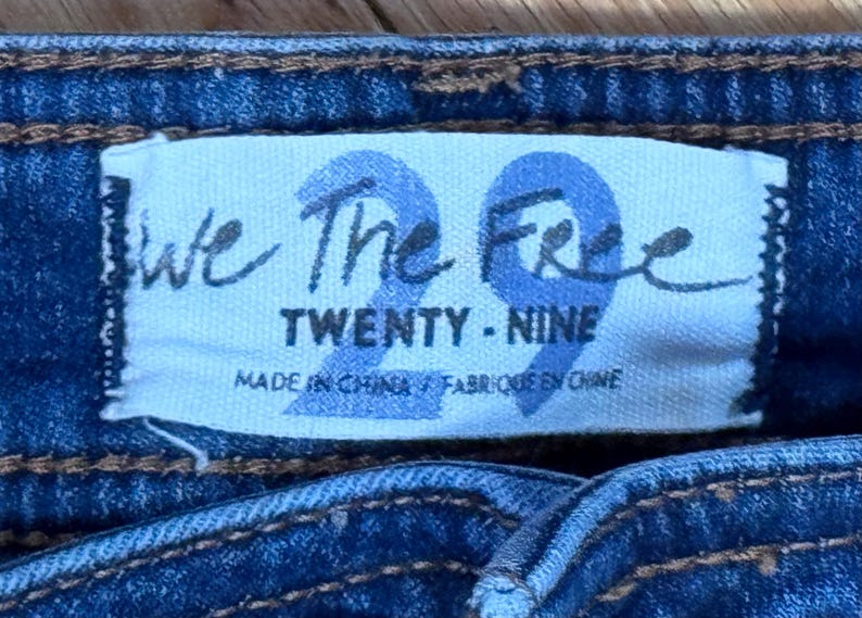 May include: Close-up of a blue denim garment with a white label. The label reads "We The Free" above the number "29" in a stylized font. Below, it says "TWENTY - NINE" and "MADE IN CHINA / FABRIQU&Eacute; EN CHINE".