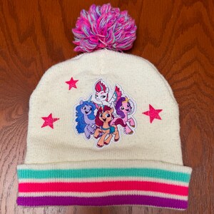 Puede incluir: Gorro de punto blanco con un pompón rosa en la parte superior. El gorro tiene un patrón de rayas de colores en la parte inferior y un parche de cinco ponis de dibujos animados en la parte delantera.