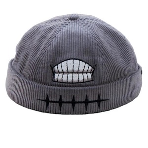May include: Gray corduroy beanie hat with an embroidered grinning mouth design. The hat features a ribbed texture and a rolled-up brim. The mouth design is white with black outlines and stitching.