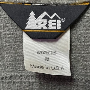 May include: A white tag with black text that reads "Womens M Made in U.S.A." attached to a black tag with a white logo that reads "REI" with a mountain and tree design.