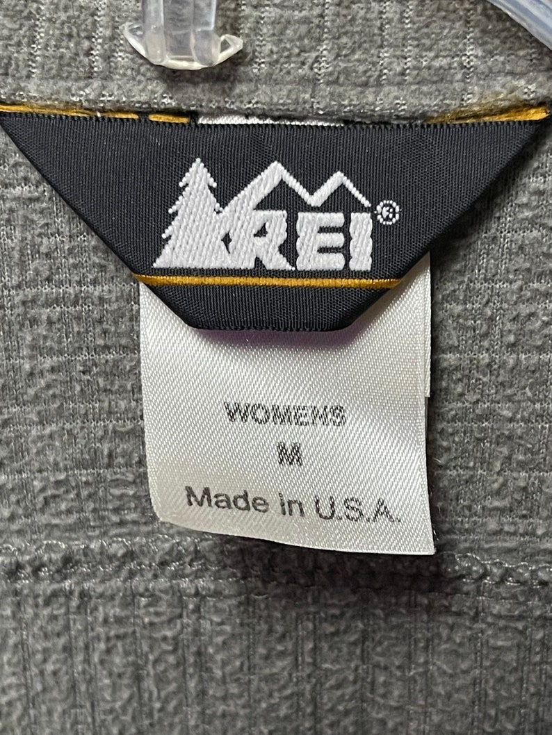 May include: A white tag with black text that reads "Womens M Made in U.S.A." attached to a black tag with a white logo that reads "REI" with a mountain and tree design.