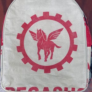 Könnte beinhalten: Ein beigefarbener Rucksack aus recycelten Materialien mit einem roten geflügelten Pferdesymbol und einem roten Zahnrad-Design. Der Rucksack hat schwarze Träger und eine Netztasche an der Seite.
