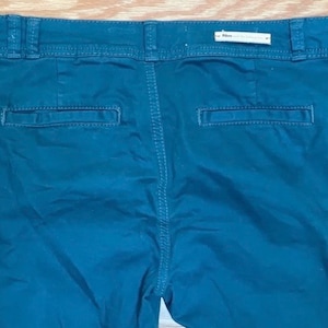 Puede incluir: Pantalones de color verde azulado oscuro con dos bolsillos traseros y una cintura. Los pantalones tienen cierre de bot&oacute;n y cremallera. La parte trasera de los pantalones es visible, mostrando las costuras y la etiqueta de la marca.