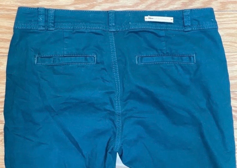 Puede incluir: Pantalones de color verde azulado oscuro con dos bolsillos traseros y una cintura. Los pantalones tienen cierre de bot&oacute;n y cremallera. La parte trasera de los pantalones es visible, mostrando las costuras y la etiqueta de la marca.