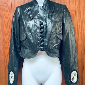 Könnte beinhalten: Eine schwarze, kurze Jacke mit Stehkragen und Knopfleiste. Die Jacke hat dekorative Knöpfe an den Schultern und einen Totenkopf- und Knochen-Patch an einem Ärmel und einen High-Heel-Schuh-Patch am anderen Ärmel.