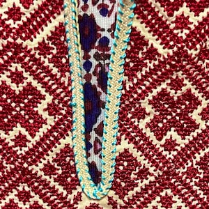 May include: Close-up of a red and white embroidered fabric with a blue and white trim. The fabric has a geometric pattern.