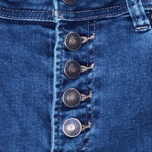 May include: Close-up of blue denim jeans with a button-up fly. The jeans have five silver-colored buttons and visible stitching details. The denim fabric is a medium wash.