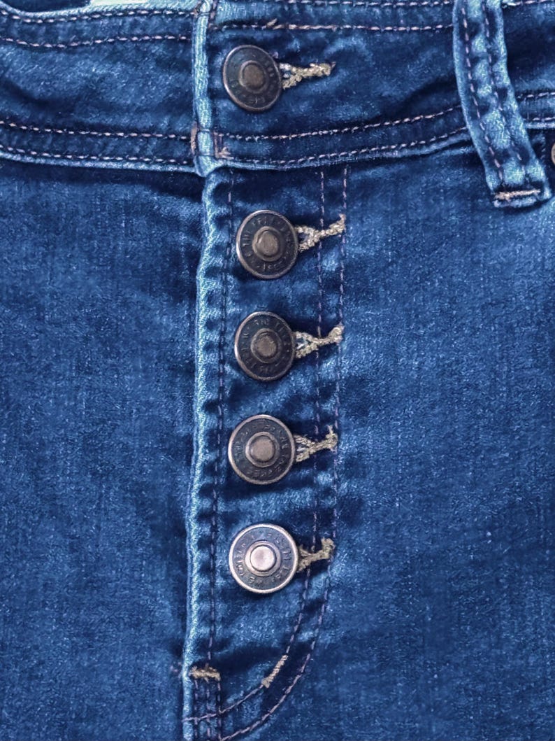 May include: Close-up of blue denim jeans with a button-up fly. The jeans have five silver-colored buttons and visible stitching details. The denim fabric is a medium wash.