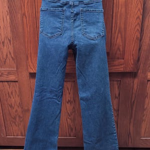 May include: Dark blue denim jeans with a high waist and flared legs. The back view shows two pockets and a waistband with a button closure. The jeans are made of a sturdy denim fabric.