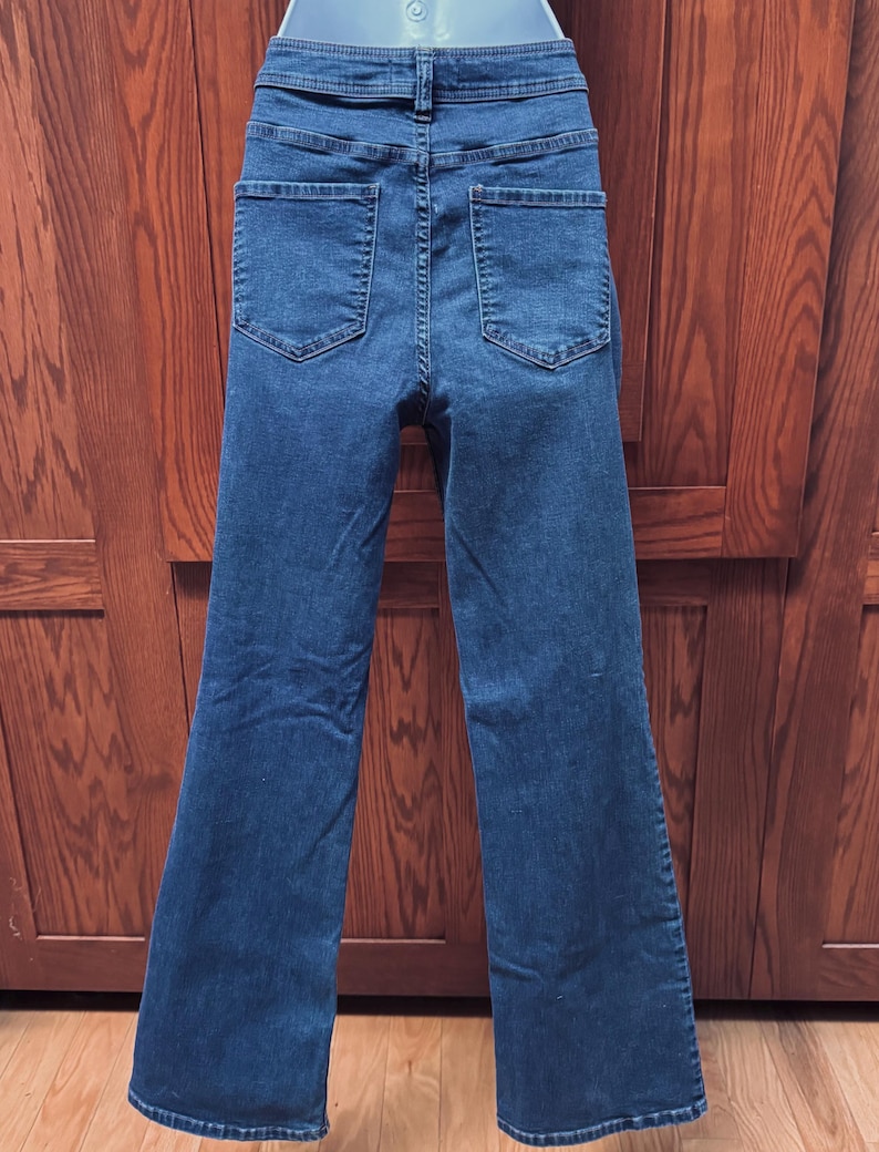 May include: Dark blue denim jeans with a high waist and flared legs. The back view shows two pockets and a waistband with a button closure. The jeans are made of a sturdy denim fabric.