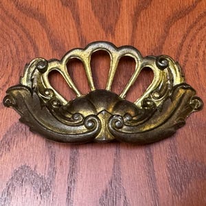 Könnte beinhalten: Verzierter antiker Schubladengriff aus Messing mit fächerförmigem Design. Der Griff hat eine zentrale, stilisierte Fächerform mit drei bogenförmigen Öffnungen, flankiert von Blatt- und Wirbeldetails. Das Messing hat eine warme, gealterte Patina.