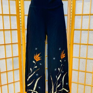 Może przedstawiać: Czarne szerokie spodnie z wysokim stanem i haftem kwiatowym na nogawkach. Haft przedstawia pomarańczowe kwiaty, białe łodygi i srebrne akcenty z brokatu.