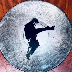 May include: A vinyl record featuring a black silhouette of a person in a dynamic pose. The record has a distressed, textured appearance with a silver and grey colour scheme. The image is set against a wooden background.