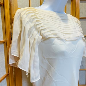 Puede incluir: Una blusa blanca de gasa con cuello plisado y mangas cortas. La blusa está hecha de una tela ligera y tiene un corte holgado.