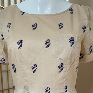 Op de afbeelding: Een beige korte mouwen jurk met een bloemenprint van kleine paarse bloemen. De jurk heeft een ronde hals en een enkele zak aan de linkerkant.