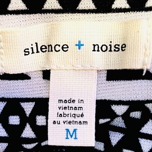 Może przedstawiać: Biała metka z czarnym tekstem, kt&oacute;ry brzmi "silence + noise" i "made in vietnam, fabriqu&eacute; au vietnam". Metka ma r&oacute;wnież niebieskie "M" oznaczające rozmiar.