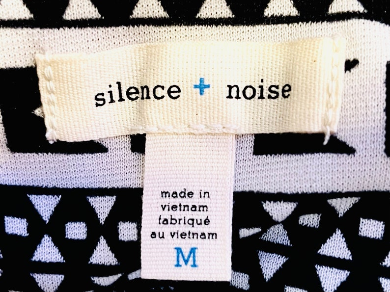 Może przedstawiać: Biała metka z czarnym tekstem, kt&oacute;ry brzmi "silence + noise" i "made in vietnam, fabriqu&eacute; au vietnam". Metka ma r&oacute;wnież niebieskie "M" oznaczające rozmiar.