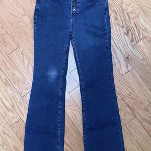 May include: Dark wash denim flare jeans with a high waist and button fly. The jeans have a classic five-pocket design and a flared leg opening. The jeans are made of a medium-weight denim fabric.