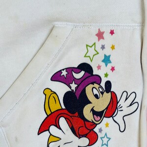 K&ouml;nnte beinhalten: Wei&szlig;er Kapuzenpullover mit einer bunten Mickey Mouse-Illustration, die einen lila Zaubererhut tr&auml;gt und auf einem gelben Stern fliegt. Das Design enth&auml;lt bunte Sterne.