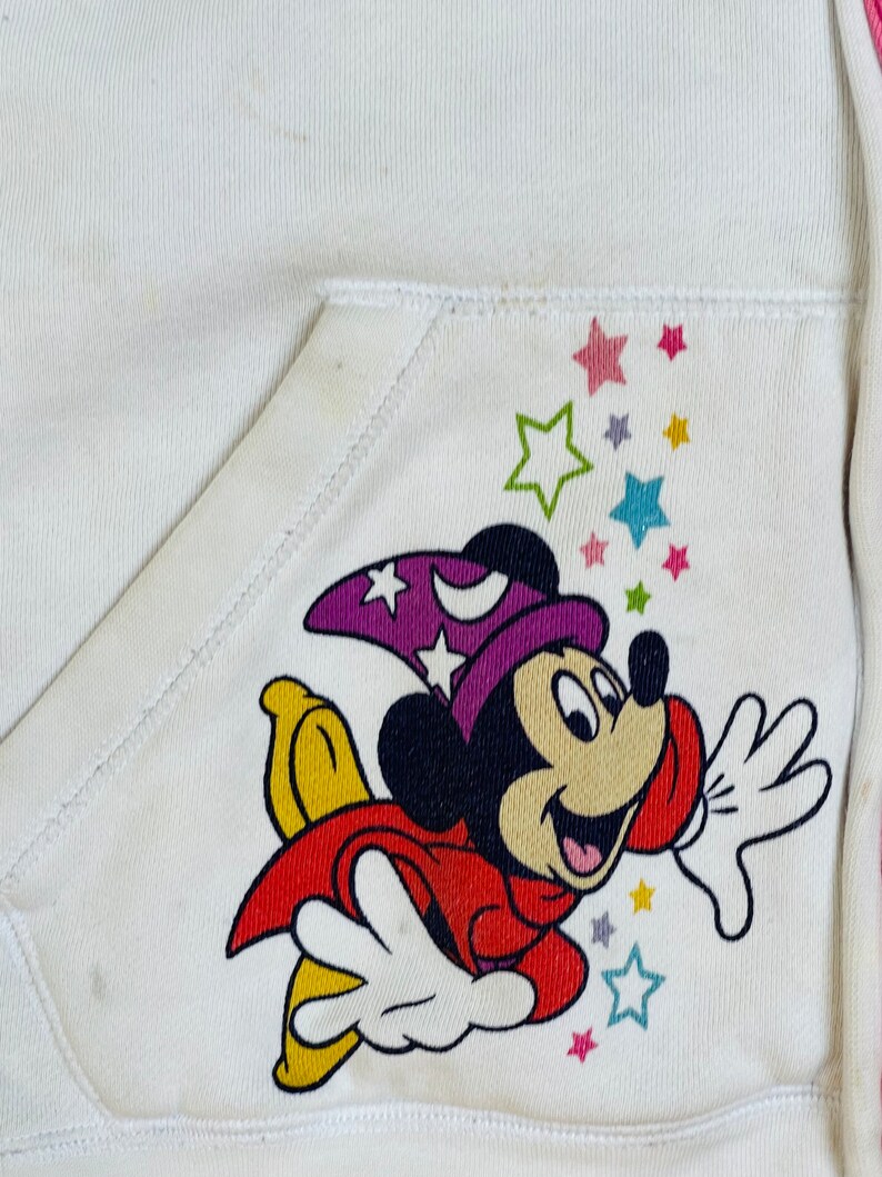 K&ouml;nnte beinhalten: Wei&szlig;er Kapuzenpullover mit einer bunten Mickey Mouse-Illustration, die einen lila Zaubererhut tr&auml;gt und auf einem gelben Stern fliegt. Das Design enth&auml;lt bunte Sterne.