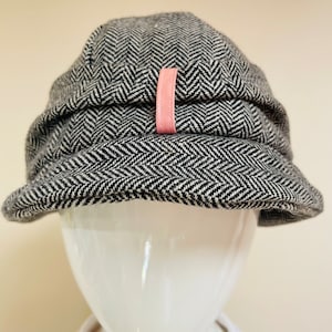 May include: A black and white herringbone patterned newsboy cap with a pink leather accent.