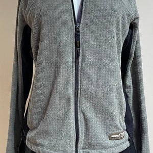 May include: A gray zippered jacket with black accents on the sleeves. The jacket has a ribbed texture and a small logo on the bottom right side.