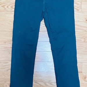 Puede incluir: Pantalones de color verde azulado oscuro con un dise&ntilde;o cl&aacute;sico de cinco bolsillos. Los pantalones tienen cierre de bot&oacute;n y cremallera. La tela es de color liso y suave. Los pantalones est&aacute;n colocados sobre una superficie de madera.