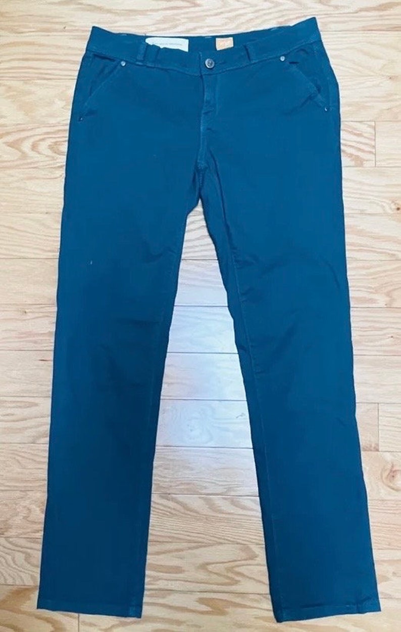Puede incluir: Pantalones de color verde azulado oscuro con un dise&ntilde;o cl&aacute;sico de cinco bolsillos. Los pantalones tienen cierre de bot&oacute;n y cremallera. La tela es de color liso y suave. Los pantalones est&aacute;n colocados sobre una superficie de madera.