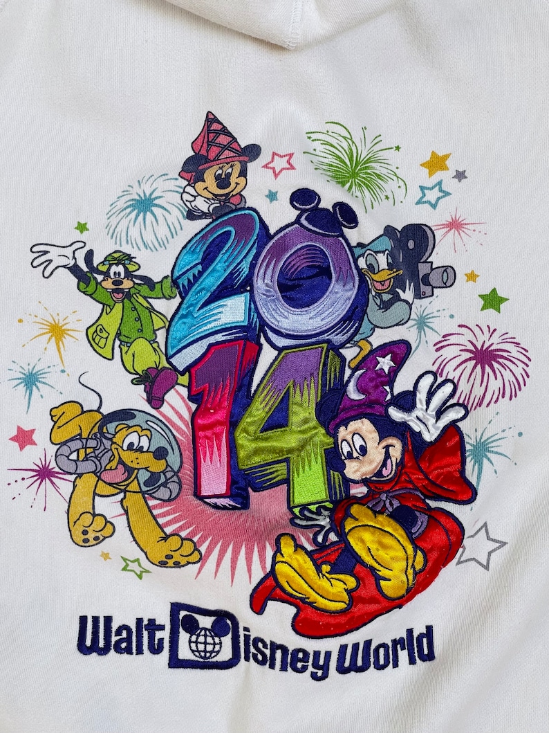 K&ouml;nnte beinhalten: Wei&szlig;er Hoodie mit einem bunten gestickten Design mit Mickey Mouse, Minnie Mouse, Goofy, Pluto und Donald Duck. Das Design beinhaltet das Jahr 2014 und Feuerwerk. Der Text "Walt Disney World" ist unter dem Design gestickt.