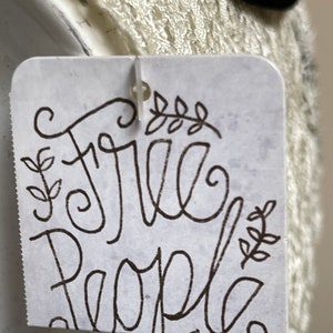 May include: A white tag with black script that reads "Free People" with a small floral design on either side of the text.