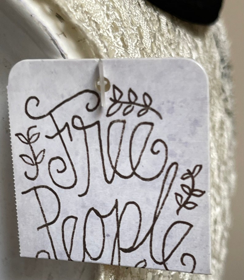 May include: A white tag with black script that reads "Free People" with a small floral design on either side of the text.