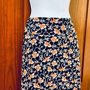 May include: A black skirt with a floral pattern of orange flowers and white vines. The skirt is displayed on a white mannequin against a wooden background. The skirt is a midi length.