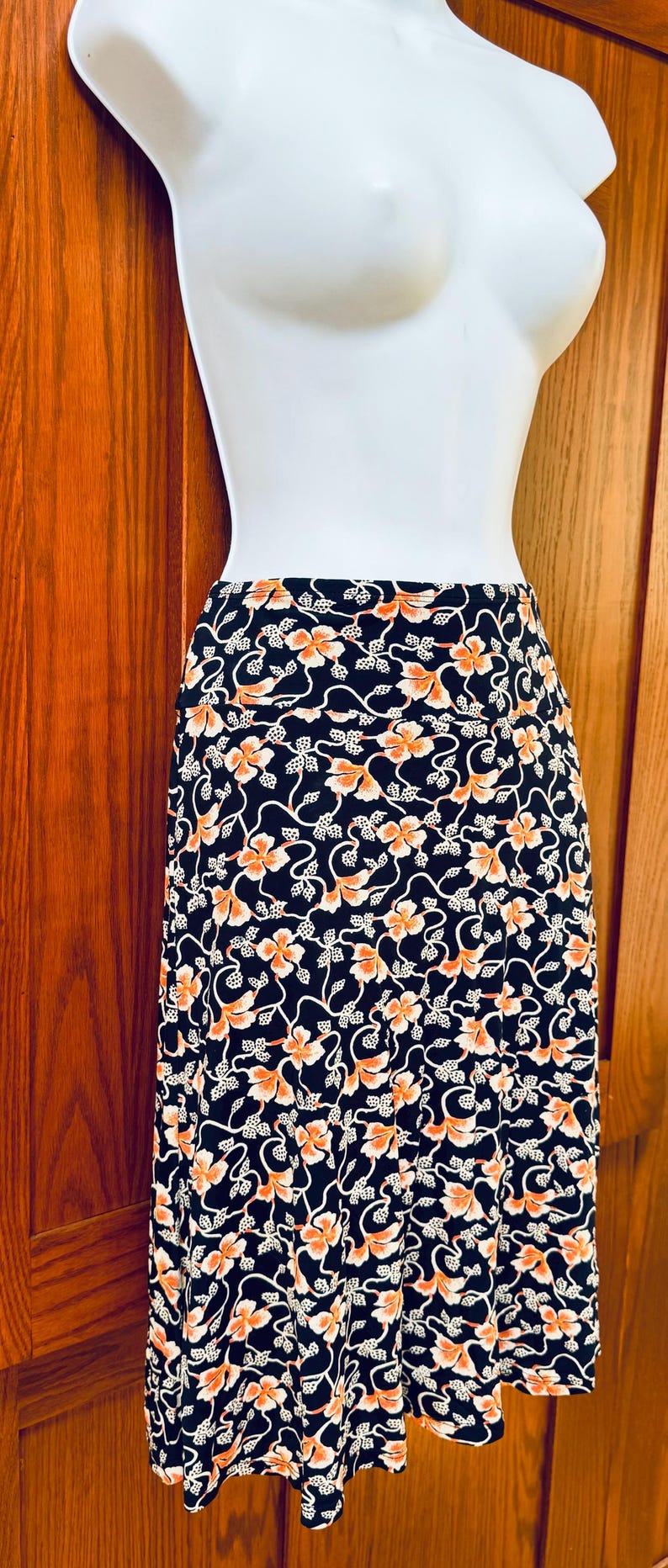 May include: A black skirt with a floral pattern of orange flowers and white vines. The skirt is displayed on a white mannequin against a wooden background. The skirt is a midi length.