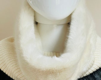 May include: A white faux fur collar on a cream-colored knit sweater. The collar is thick and fluffy, creating a luxurious look. The sweater is displayed on a white mannequin head, with a black textured garment visible below.