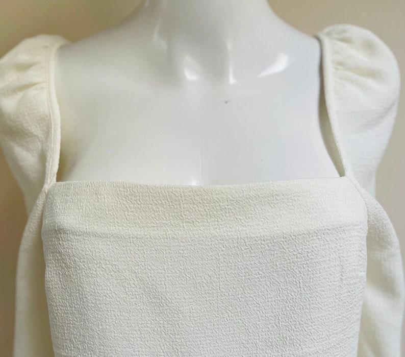 May include: A white, square-neck top with puffed sleeves. The top is made of a textured fabric.