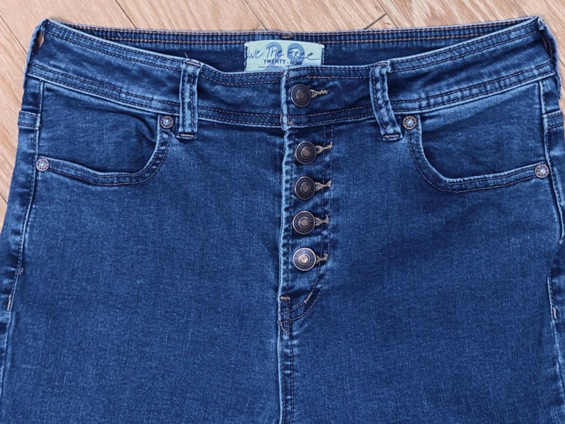 May include: Close-up of dark blue denim jeans with a button fly. The jeans have a high waist and pockets on the front and back. The brand name "We The Free" is visible on the waistband.