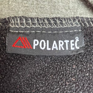 May include: A black label with white stitching and the text "POLARTEC" in white letters. A red triangle logo is on the left side of the label.