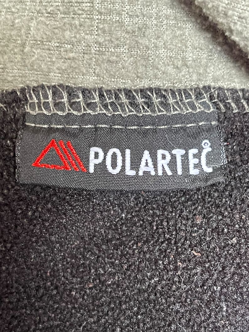 May include: A black label with white stitching and the text "POLARTEC" in white letters. A red triangle logo is on the left side of the label.
