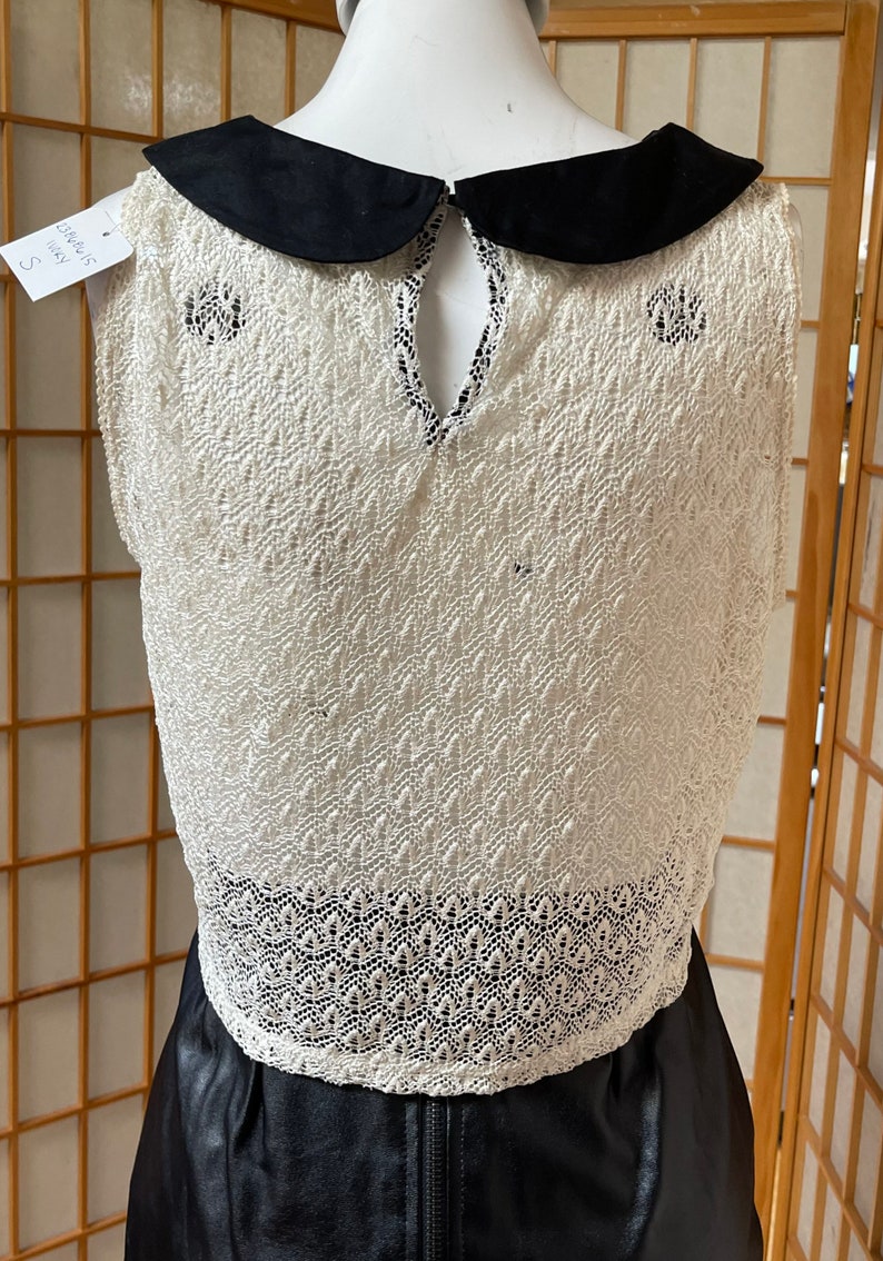 May include: White crochet sleeveless dress with a black Peter Pan collar and a black leather skirt. The dress has a lace pattern and a keyhole back.