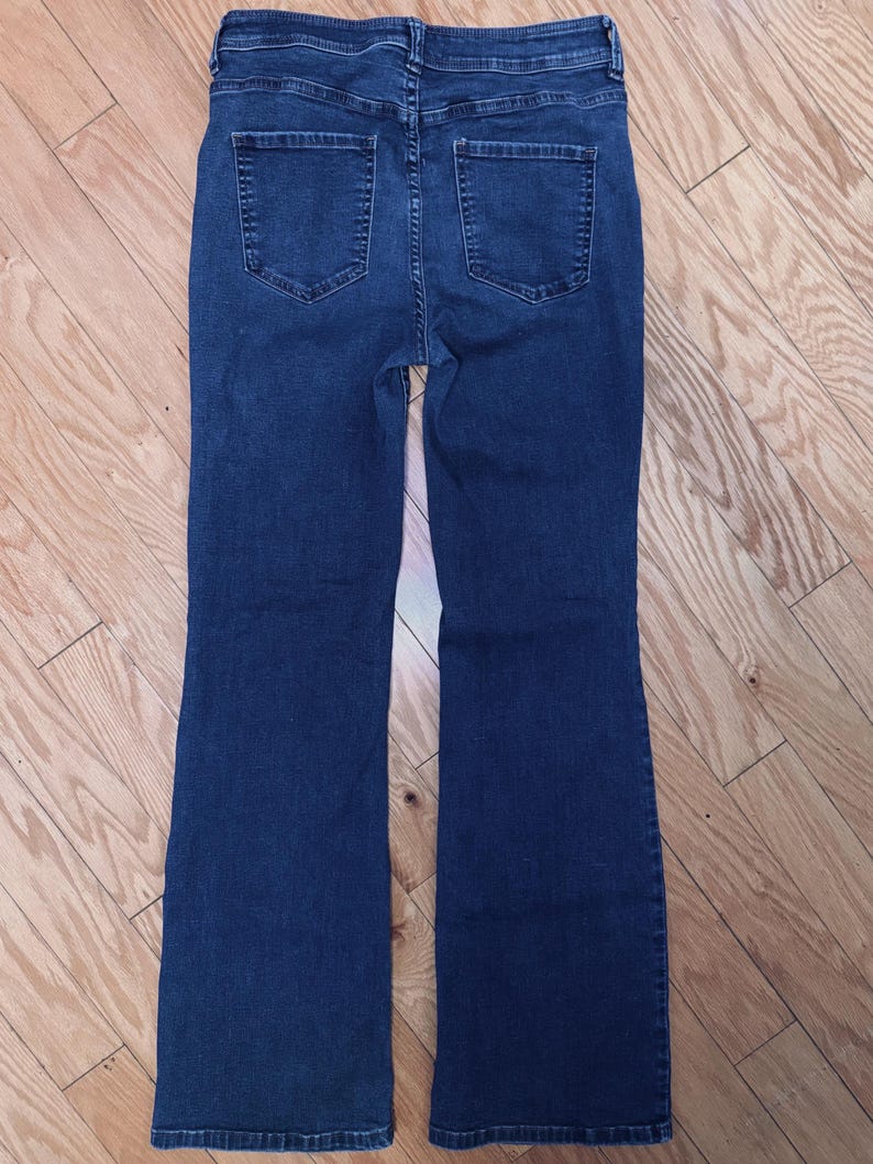 May include: Dark blue denim jeans with a high waist and flared leg. The back view shows two pockets and a classic jean design. The jeans are made of a sturdy denim material and are suitable for everyday wear.