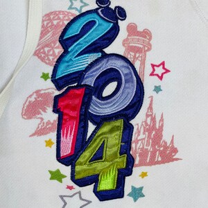 K&ouml;nnte beinhalten: Wei&szlig;er Pullover mit einem bunten gestickten Design mit den Zahlen 2014, Sternen und einer Silhouette einer Burg.