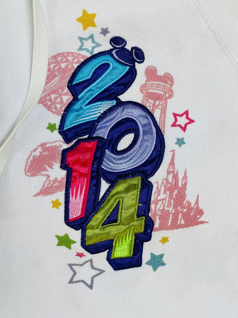 K&ouml;nnte beinhalten: Wei&szlig;er Pullover mit einem bunten gestickten Design mit den Zahlen 2014, Sternen und einer Silhouette einer Burg.