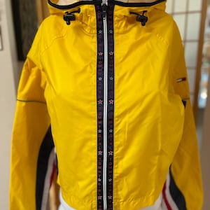 May include: Yellow hooded jacket with a black zipper and black trim. The jacket has a white and blue stripe on the sleeves and a repeating "Tommy Girl" pattern on the zipper.