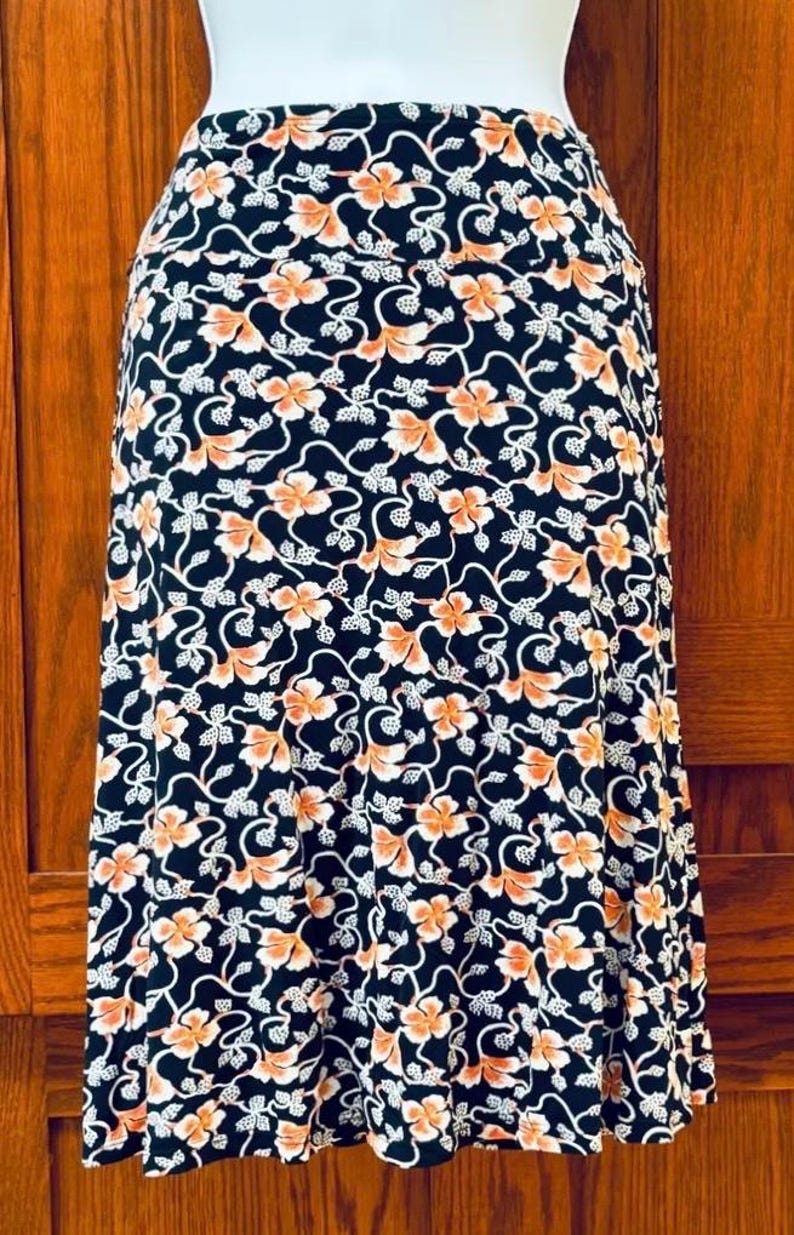 May include: A black skirt with a floral pattern. The design features orange flowers and white vines against a black background. The skirt is knee-length and made of a soft, flowing fabric.