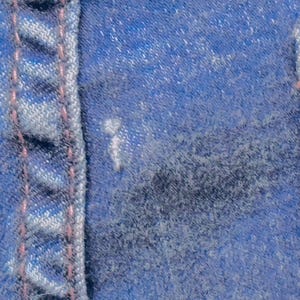 May include: Close-up of blue denim fabric with visible stitching in a lighter blue color and pink thread. The texture of the denim is rough and worn, with a few light spots.