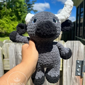 May include: A gray and black crocheted stuffed bull with white horns. The bull has black eyes and a black nose. The bull is being held in a person's hand.