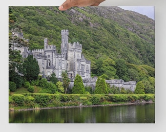 May include: A gray stone abbey with a green grassy lawn and a lake in front of it. The abbey is surrounded by lush green trees and a mountain in the background.