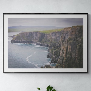 May include: A framed photograph of a dramatic coastal landscape featuring towering cliffs and a vast ocean with whitecaps. The cliffs are a mix of green grass and gray rock, with the ocean stretching out in the distance.