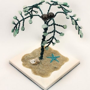 Könnte beinhalten: Ein Drahtpalmenbaum mit grünen Glasperlen für die Blätter und braunen Perlen für Kokosnüsse. Der Baum steht auf einem weißen quadratischen Sockel mit einer braunen sandartigen Oberfläche und einem blauen Seestern.