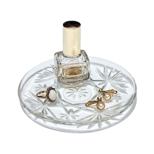 May include: A clear, round glass tray with a starburst pattern holds a small perfume bottle, a gold ring with a white stone, and a pair of gold earrings with pearls. The perfume bottle has a gold cap. The tray is a decorative item.